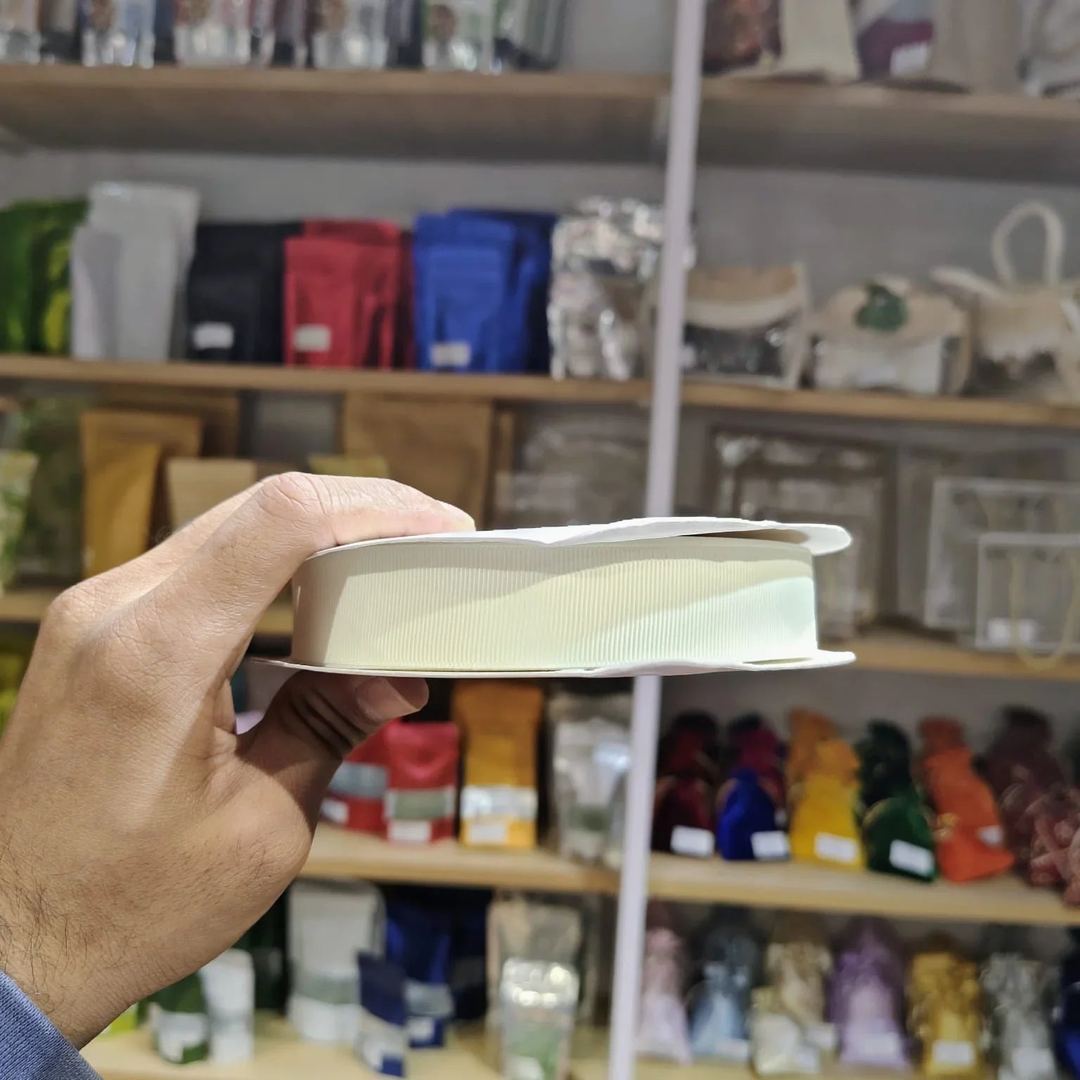 A hand holding a white ribbed rectangular item, likely a small box or tray, in front of a blurred store shelf background filled with colorful packaged goods.