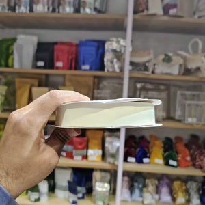 A hand holding a white ribbed rectangular item, likely a small box or tray, in front of a blurred store shelf background filled with colorful packaged goods.