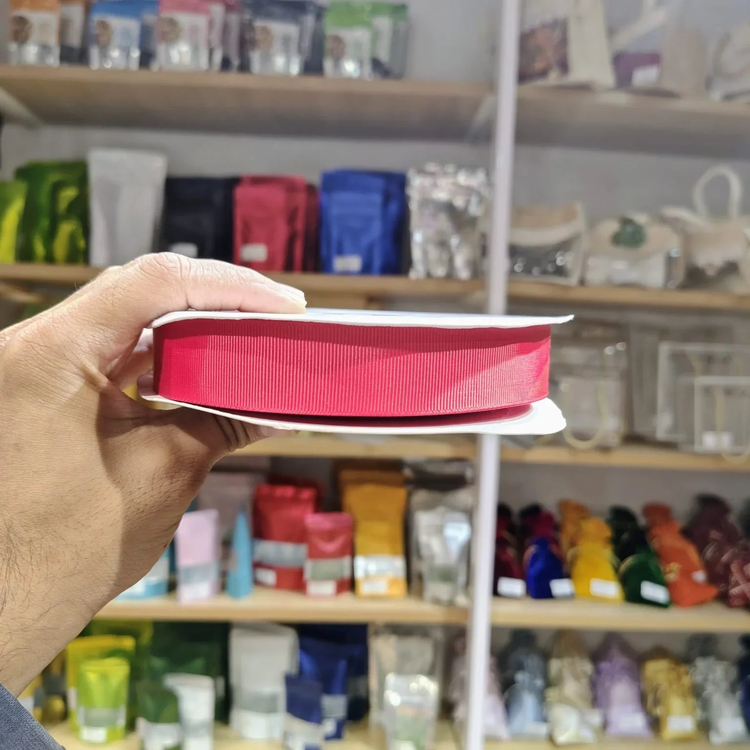 A hand holding a spool of bright pink ribbon in front of a blurred store shelf background filled with colorful packaged items.