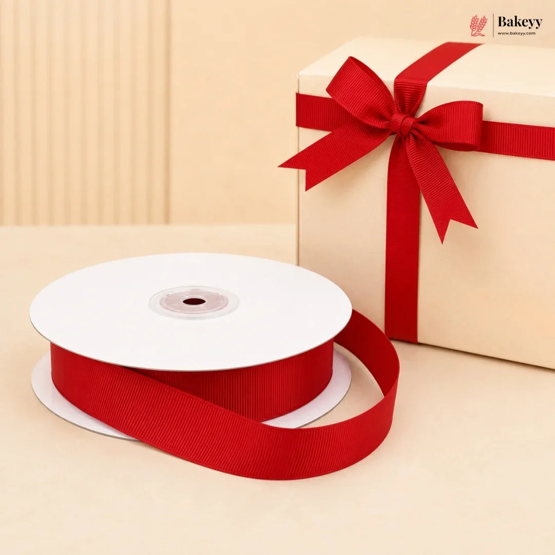A spool of red grosgrain ribbon with a matching red gift box tied in a bow, set against a warm beige background. The Bakeyy logo is visible in the top right corner.