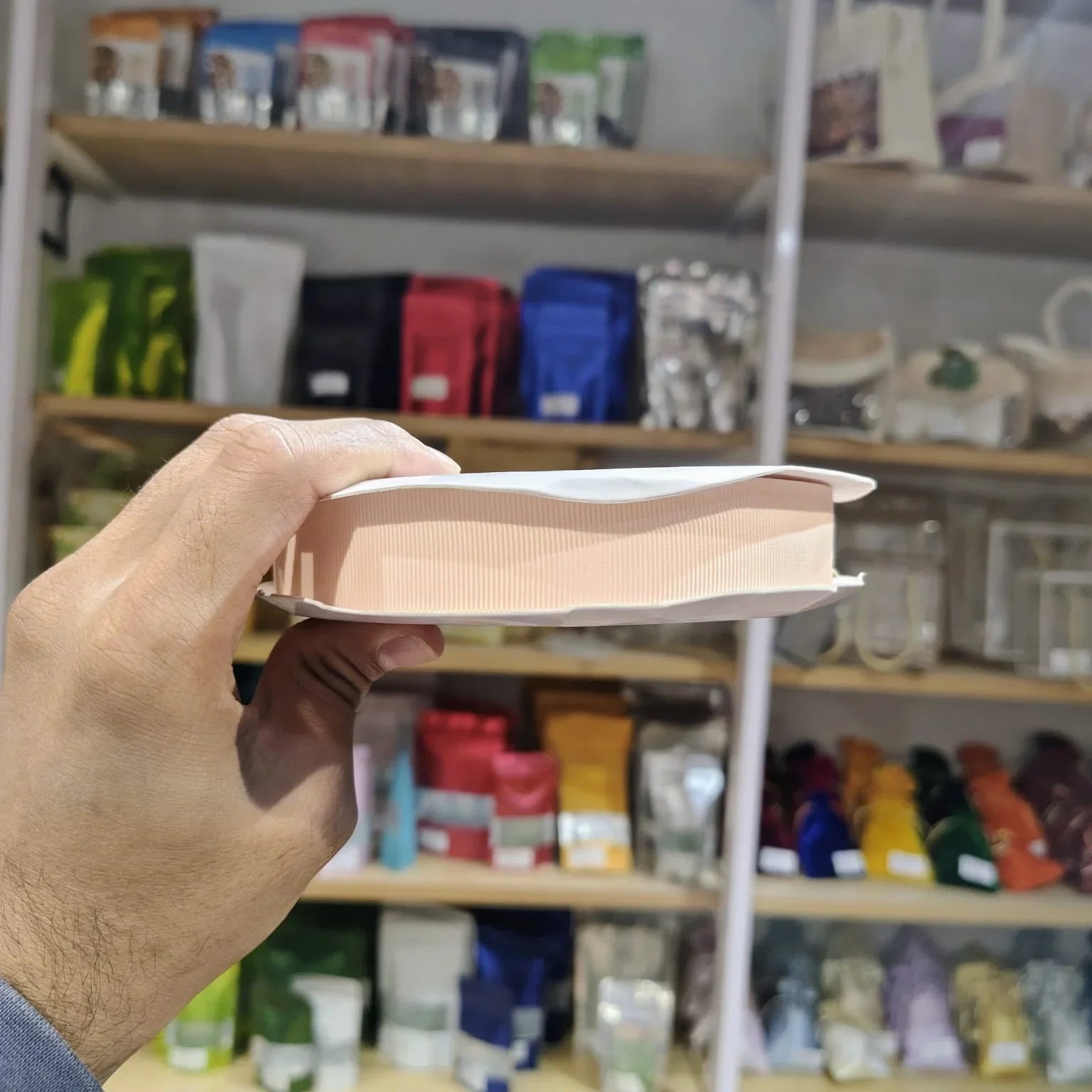 A hand holding a light peach-colored rectangular product, likely a book or a small box, in front of a blurred store shelf filled with colorful packaged items.
