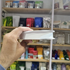 A hand holding a white rectangular product, likely a book or a slim box, in front of blurred shelves filled with colorful packaged items.