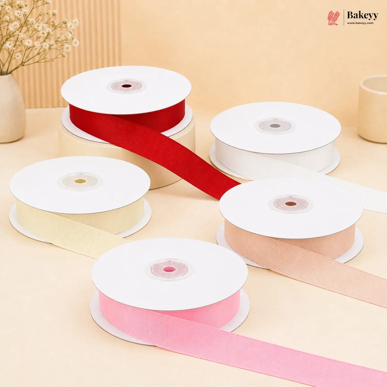 Five spools of ribbon in red, cream, white, pink, and peach, with some ribbons unspooled, arranged on a beige surface with a decorative dried flower vase in the background. The Bakeyy logo is visible in the top right corner.