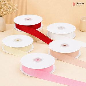 Five spools of ribbon in red, cream, white, pink, and peach, with some ribbons unspooled, arranged on a beige surface with a decorative dried flower vase in the background. The Bakeyy logo is visible in the top right corner.