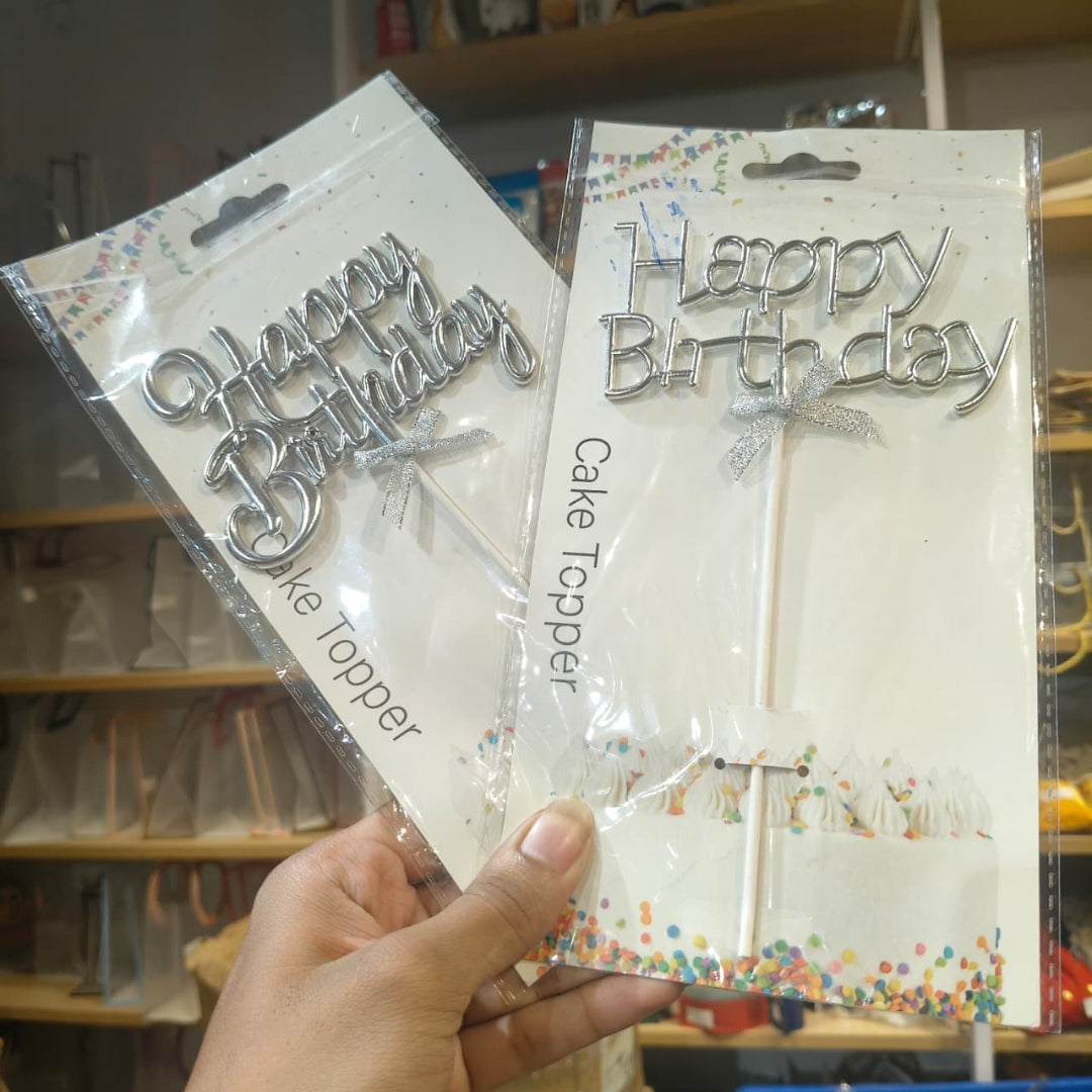 Two packaged silver 'Happy Birthday' cake toppers held in a hand, with shelves of similar items in the background.