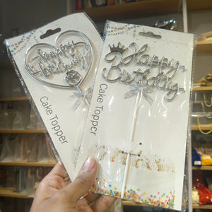 A hand holds two packaged silver metal cake toppers in a store. One topper is a heart shape with 'Happy Birthday' and a rose, and the other is a script 'Happy Birthday' with a crown and bow.