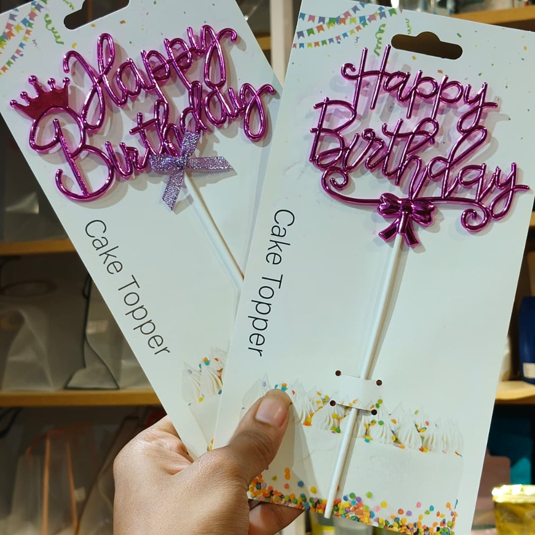 Two packaged metallic pink 'Happy Birthday' cake toppers with small bows and a crown, held in a hand against a blurred store background.