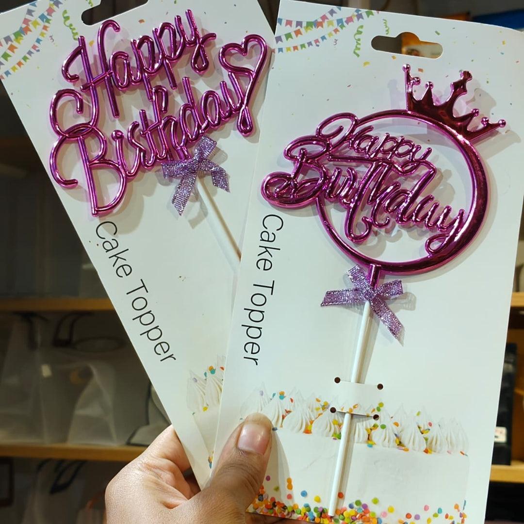 Two pink metallic 'Happy Birthday' cake toppers on white backing cards, held by a hand. One topper has a heart outline and the other has a circular lollipop shape with a small crown on top.