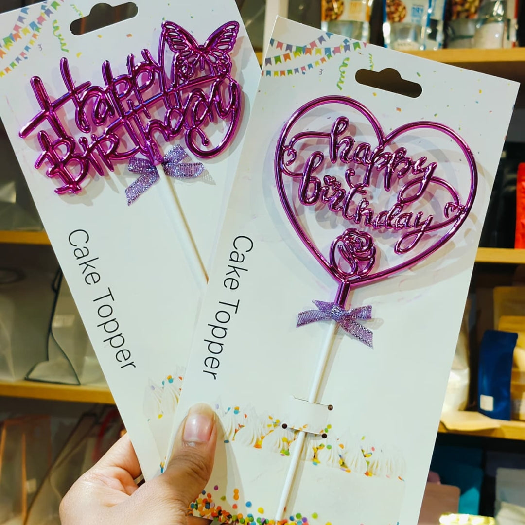 Two packaged purple glitter cake toppers on white cards, held in a hand. One topper has a butterfly and the other has a heart outline, both with 'Happy Birthday' script and small purple ribbon bows.