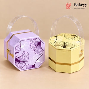 Two octagonal bakery gift boxes with clear plastic handles, shown on a beige background. The left box is lavender purple with purple ginkgo leaf patterns and gold trim.