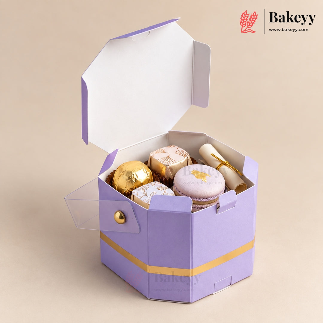 A lavender and gold hexagonal cardboard gift box with a clear hinged lid, shown open and filled with assorted pastries including macarons and chocolate-covered spheres. The Bakeyy brand logo is visible in the top right corner.