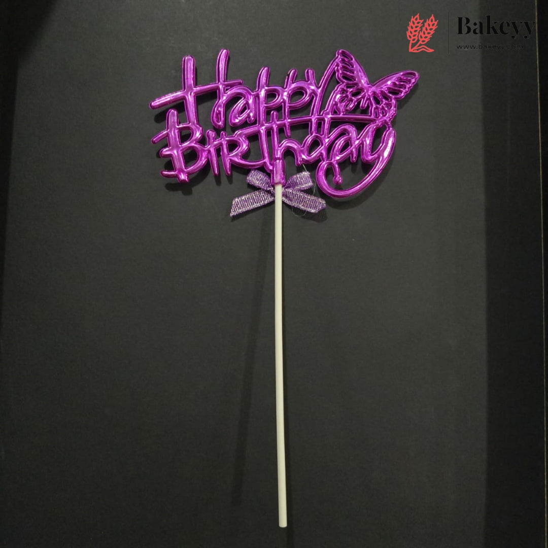 A purple glittery 'Happy Birthday' cake topper with a butterfly design and a small bow, mounted on a white stick, set against a dark background.