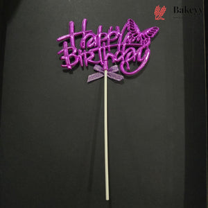 A purple glittery 'Happy Birthday' cake topper with a butterfly design and a small bow, mounted on a white stick, set against a dark background.