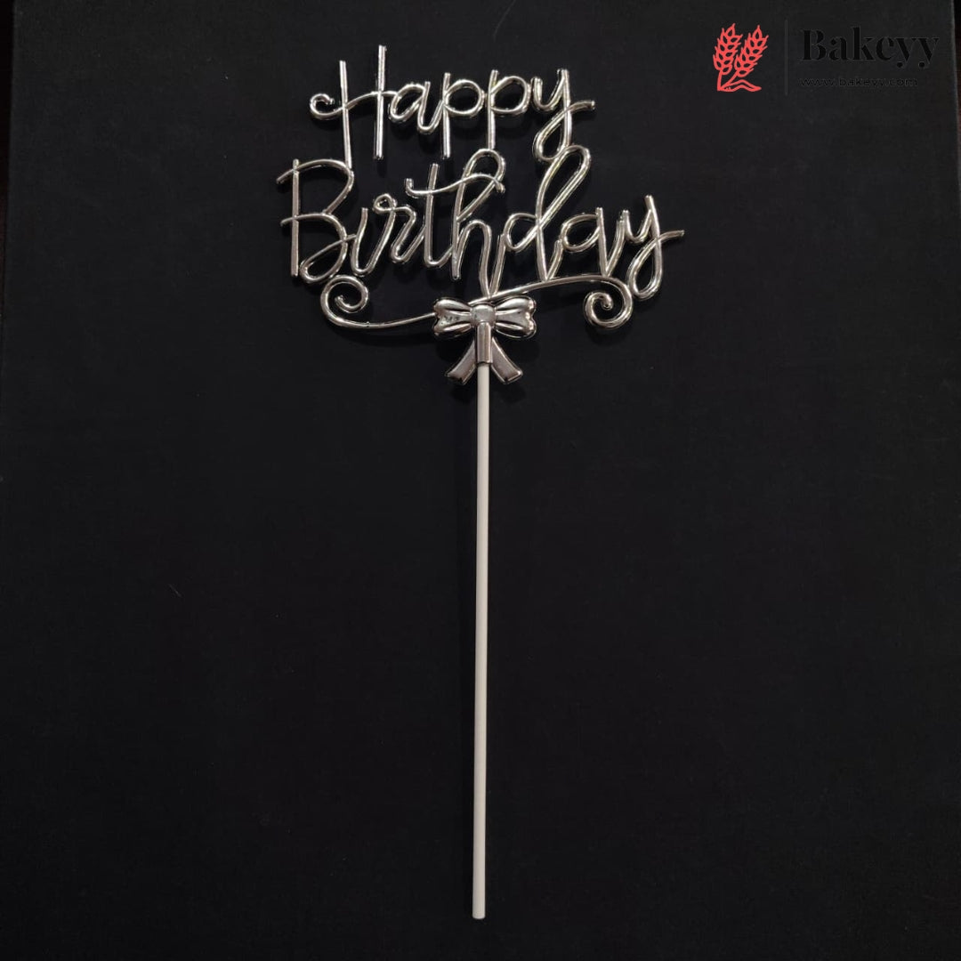 A silver metal 'Happy Birthday' cake topper with a bow detail and long stick, displayed against a solid black background. A Bakey bakery logo is visible in the top right corner.