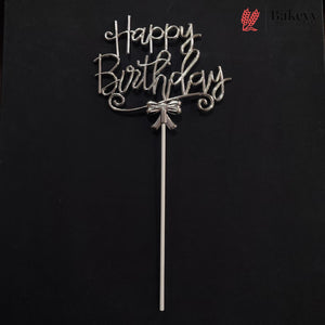 A silver metal 'Happy Birthday' cake topper with a bow detail and long stick, displayed against a solid black background. A Bakey bakery logo is visible in the top right corner.