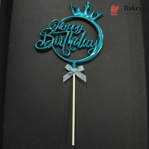 A metallic turquoise blue 'Happy Birthday' cake topper with a crown on a stick, displayed against a dark background. A Bakeyy logo is visible in the top right corner.