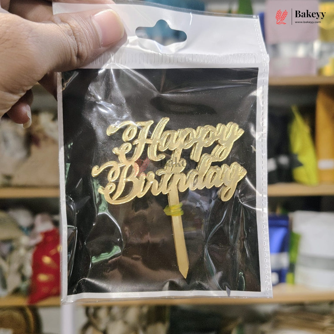 Gold 'Happy Birthday' cake topper in clear packaging held by a hand, with a blurred background.