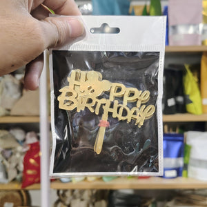 Gold 'Happy Birthday' cake topper in packaging held by a hand with a blurred store background.