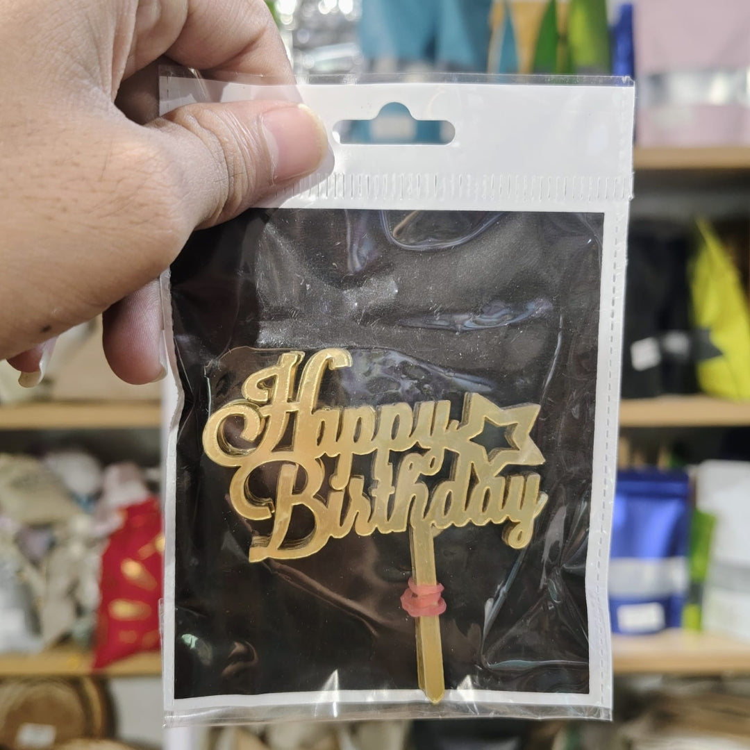 Gold 'Happy Birthday' cake topper in packaging held by a hand with a blurred store background.