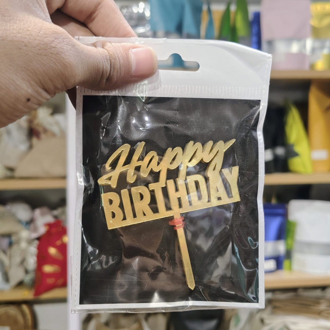 Hand holding a package with a 'Happy Birthday' decoration inside, against a blurred store background.