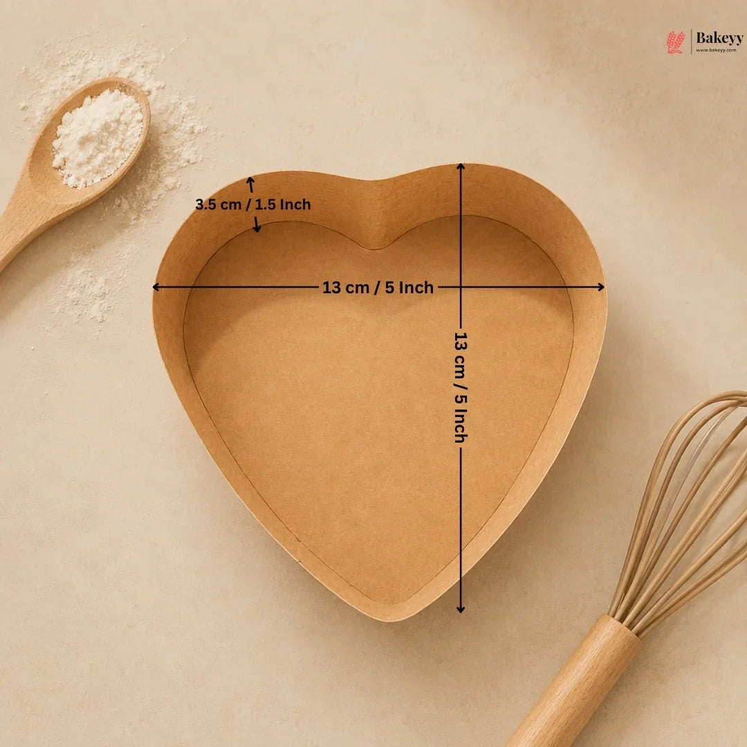 Heart-shaped wooden baking dish with measurements on a beige background, accompanied by a wooden spoon and whisk.