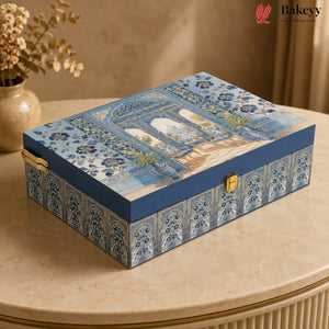 A rectangular decorative gift box with a blue and white architectural pattern on the lid and sides, featuring gold-tone latches, placed on a beige marble table in a warm indoor setting.