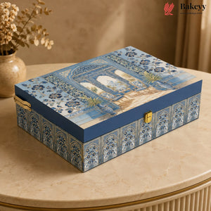 A rectangular decorative gift box with a blue and white architectural pattern on the lid and sides, featuring gold-tone latches, placed on a beige marble table in a warm indoor setting.