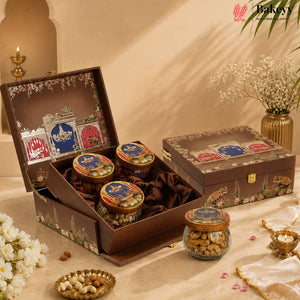 An open and closed decorative gift box containing three glass jars of nuts, arranged on a beige surface with flowers, candles, and a warm neutral background. The Bakey brand logo appears in the top right corner.