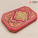 An ornate rectangular cake box with a vibrant pink lid decorated with intricate gold, green, and black traditional patterns, shown on a neutral beige background.