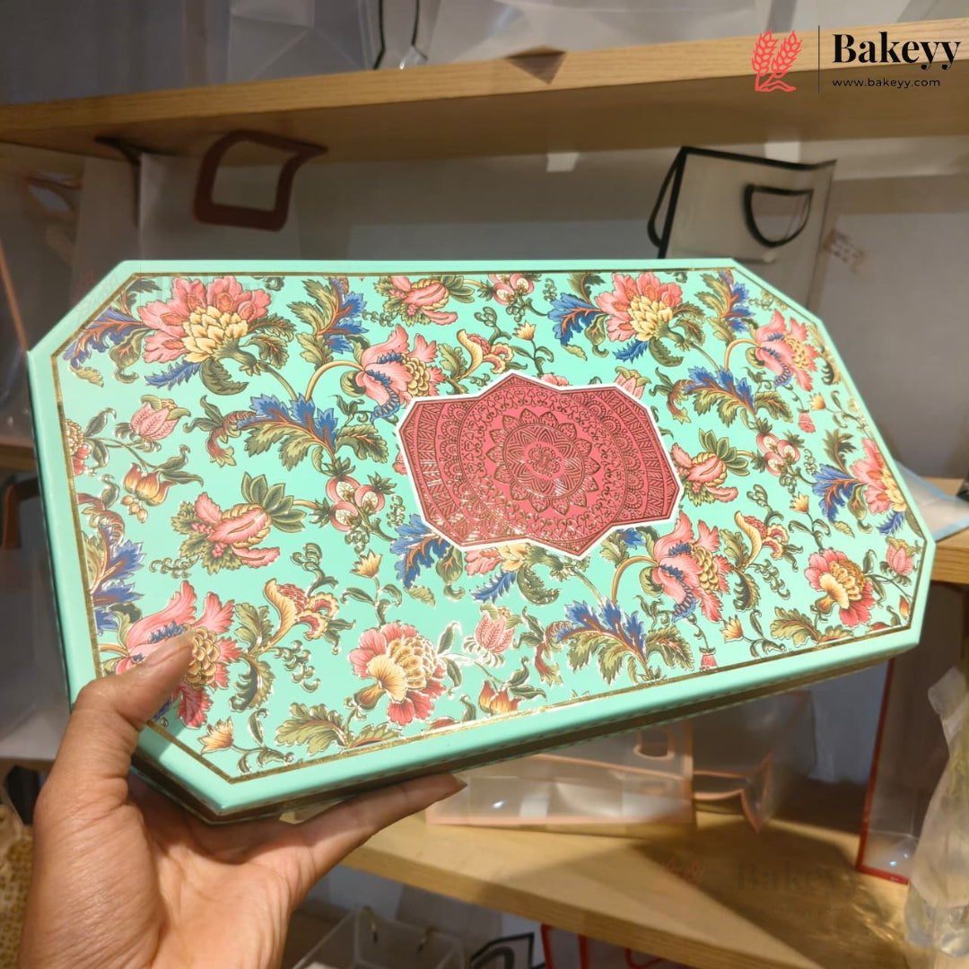 A hand holds a decorative octagonal mint green tin box with a colorful floral pattern and a red central medallion. The background shows a bakery shelf with bags and products.