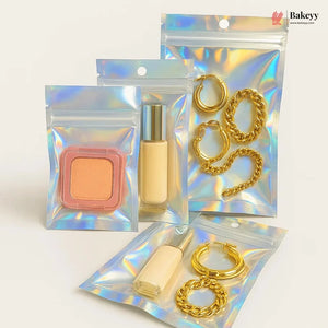 Four iridescent holographic zipper pouches arranged on a light beige background. Two pouches contain gold-tone chain and hoop earrings, and two contain cosmetic items: a peach-colored square compact and a beige liquid bottle with a silver cap.