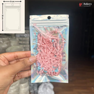 A hand holds a clear holographic zip-top pouch containing pink shredded paper filler. The pouch has a printed measurement diagram in the top-left corner showing 9 cm by 15 cm dimensions, and a Bakeyy logo watermark appears in the top-right.
