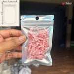 A hand holds a small holographic zip-top pouch filled with pink shredded paper against a dark indoor background. A white inset in the top left shows a simple packaging diagram labeled 7.