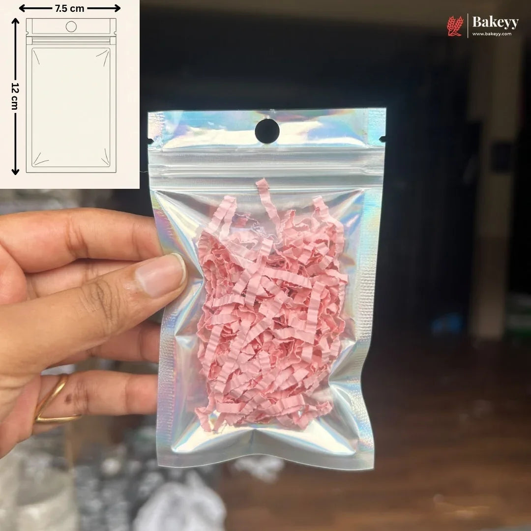 A hand holds a small holographic zip-top pouch filled with pink shredded paper against a dark indoor background. A white inset in the top left shows a simple packaging diagram labeled 7.