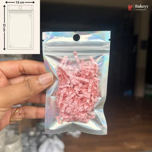 A hand holds a small holographic zip-top pouch filled with pink shredded paper against a dark indoor background. A white inset in the top left shows a simple packaging diagram labeled 7.