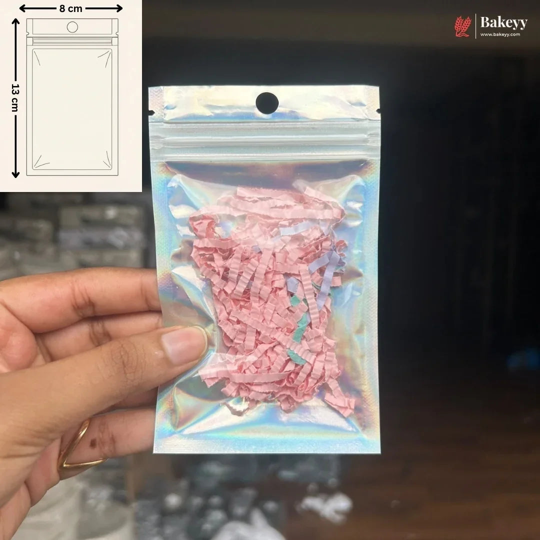 A hand holds a small holographic zipper pouch containing pink shredded paper filler. The pouch has a die-cut hanging hole at the top.