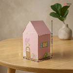Pink house-shaped box with measurements on a wooden table, featuring a plant in the background.