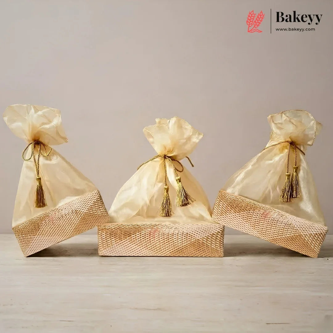 Three gold satin drawstring gift bags with tassel ties, arranged on top of a woven gold box against a neutral beige background. The Bakeyy logo is visible in the top right corner.