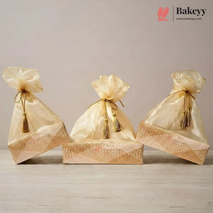 Three gold satin drawstring gift bags with tassel ties, arranged on top of a woven gold box against a neutral beige background. The Bakeyy logo is visible in the top right corner.