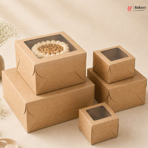A set of kraft brown bakery boxes with clear window lids, shown in multiple sizes. One larger box is stacked on top of another, holding a round cake with white frosting and nuts.