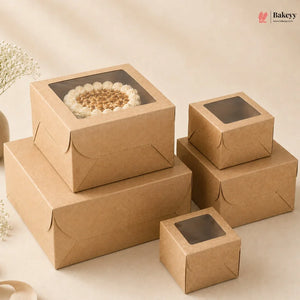 A set of kraft brown bakery boxes with clear window lids, shown in multiple sizes. One larger box is stacked on top of another, holding a round cake with white frosting and nuts.