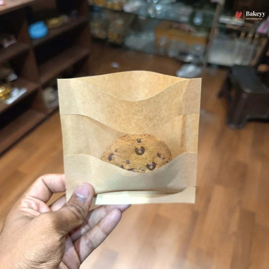Hand holding a brown paper bag with a chocolate chip cookie inside, blurred store interior in the background.