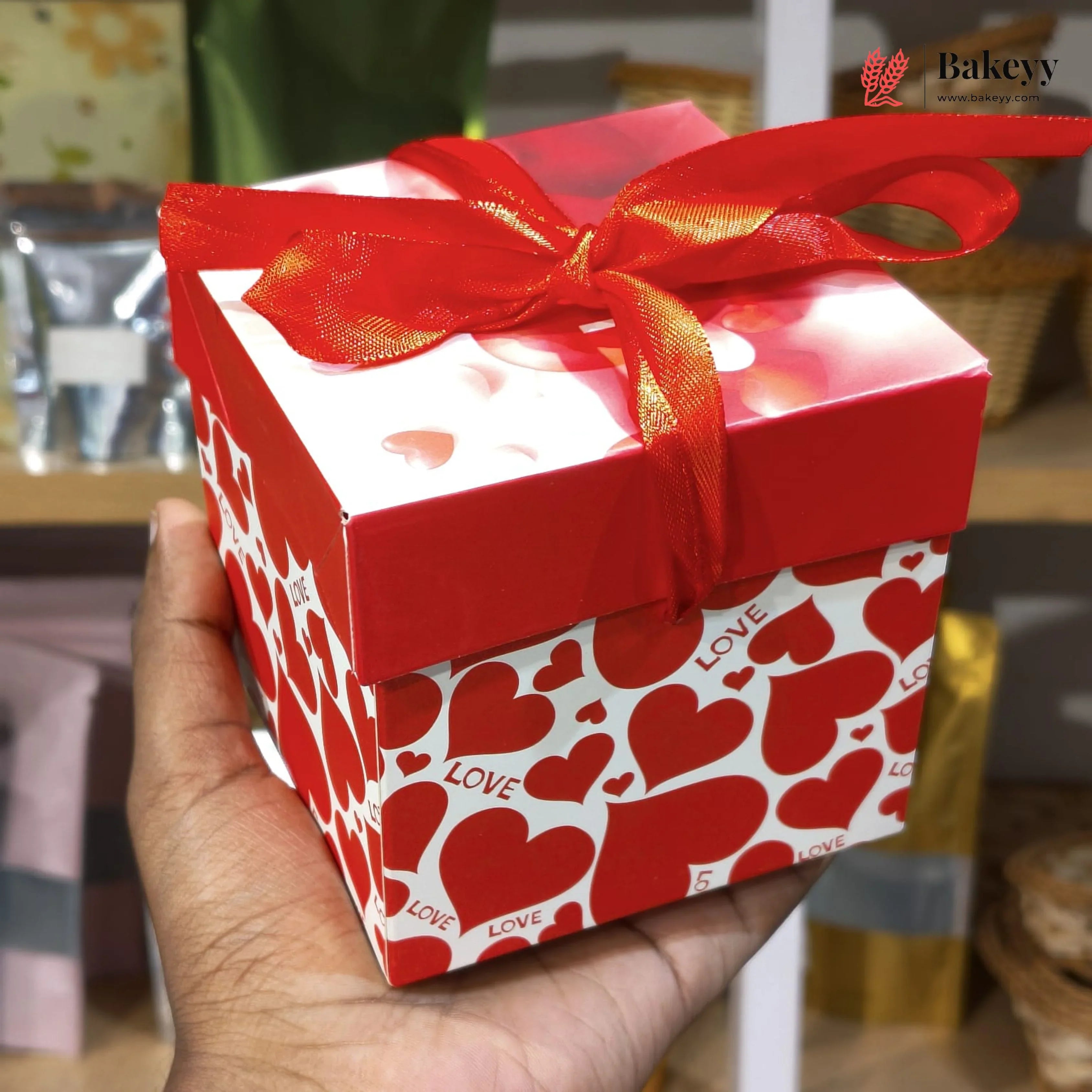 Red gift box with heart patterns and a red ribbon held by a hand, with 'Bakeyy' branding in the corner.