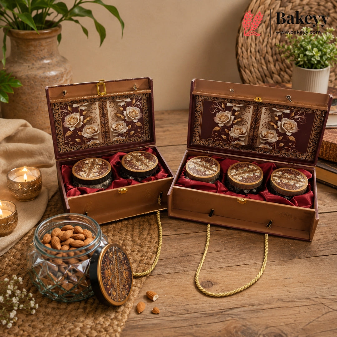 Two ornate brown wooden gift boxes with floral inlay and red velvet lining, each holding three round jars, displayed on a wooden table with candles, a glass jar of almonds, and decorative props. The Bakey brand logo appears in the top right corner.