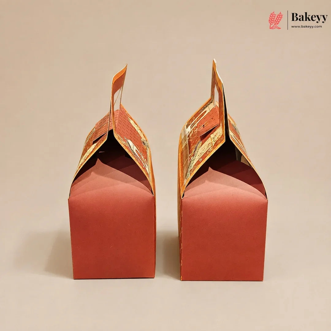 Two red paper gift boxes with orange printed handles, shown from the back on a plain beige background. A Bakeyy logo is visible in the top right corner.