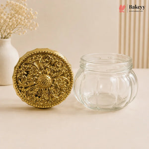 An empty clear glass jar with a decorative round gold lid placed beside it on a beige surface, with a vase of dried flowers and a vertical ribbed panel in the background.