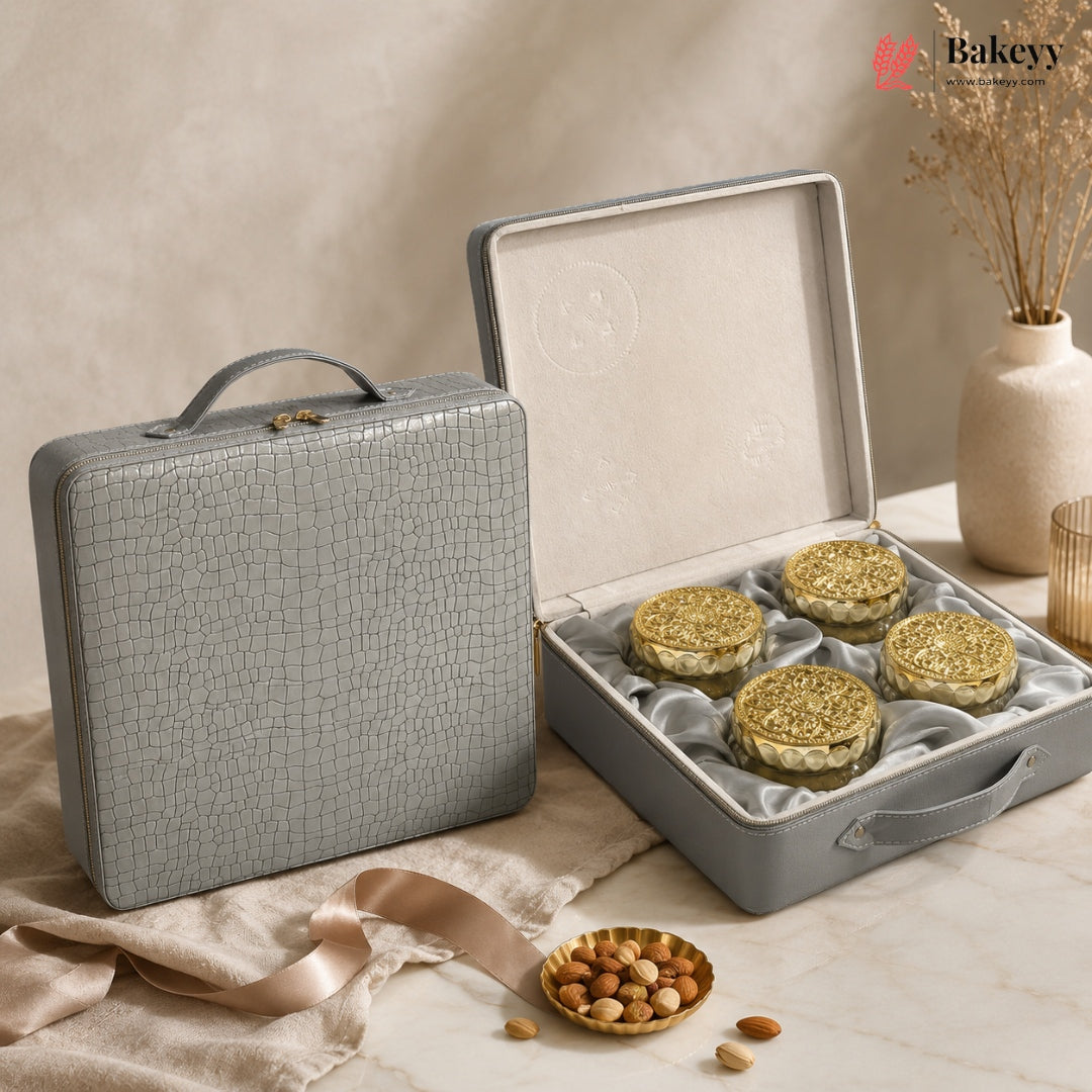 A gray textured hard case shown closed beside an open case displaying four round gold-lidded pastries or cookies on a cream satin insert. The scene is styled on a marble surface with beige fabric, a small dish of nuts, and dried flowers in a vase.