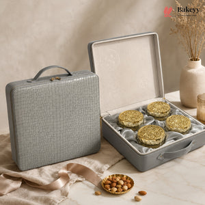 A gray textured hard case shown closed beside an open case displaying four round gold-lidded pastries or cookies on a cream satin insert. The scene is styled on a marble surface with beige fabric, a small dish of nuts, and dried flowers in a vase.