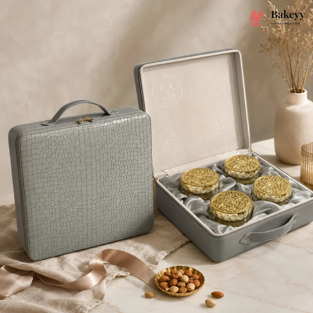 A gray textured hard case shown closed beside an open case displaying four round gold-lidded pastries or cookies on a cream satin insert. The scene is styled on a marble surface with beige fabric, a small dish of nuts, and dried flowers in a vase.