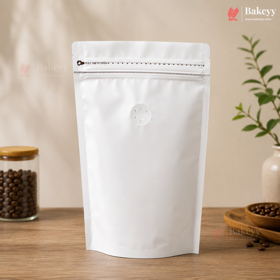 A blank white stand-up resealable pouch with a zipper closure and a small circular vent hole, standing on a wooden surface. The background is a warm beige wall with soft shadows, and decorative props including a jar of coffee beans and a small plant are visible on both sides.